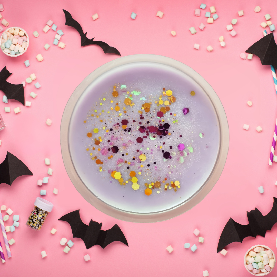 Purple wax melt with colorful confetti in a white cup on a pink background with Halloween decorations.