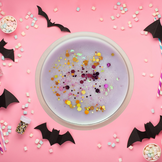Purple wax melt with colorful confetti in a white cup on a pink background with Halloween decorations.